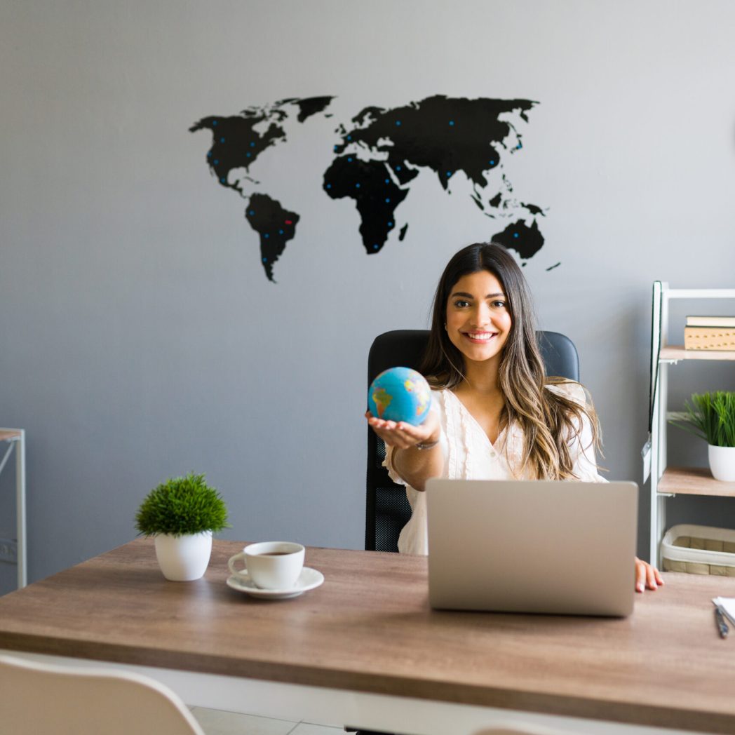 Holding the world in my hands. Attractive female travel agent showing a globe and smiling while working at her modern office